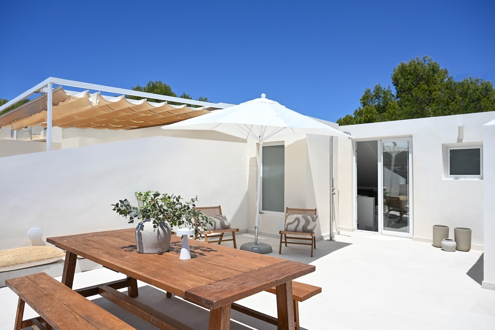 Terraza blanca soleada con mesa de comedor de madera, sombrilla y tumbonas bajo un cielo azul despejado.