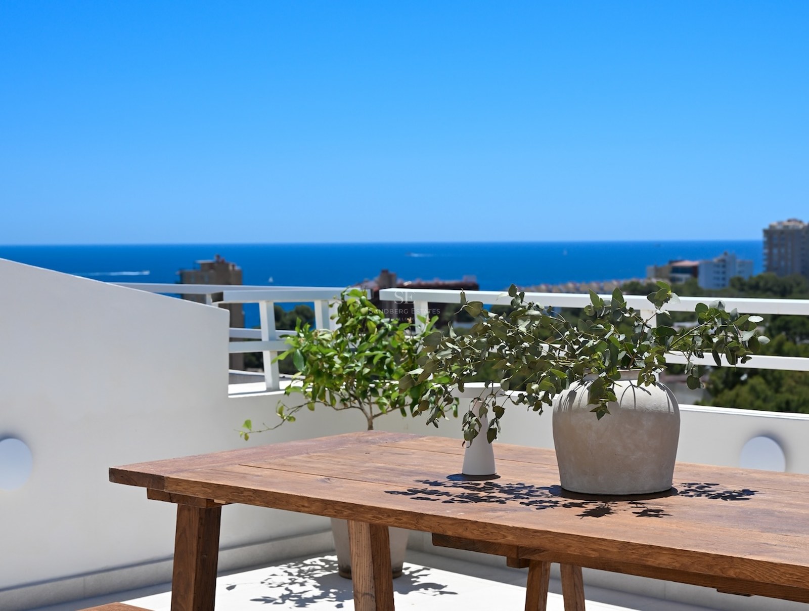 Mesa de madera rústica en una terraza blanca con vistas al mar azul profundo bajo un cielo despejado.