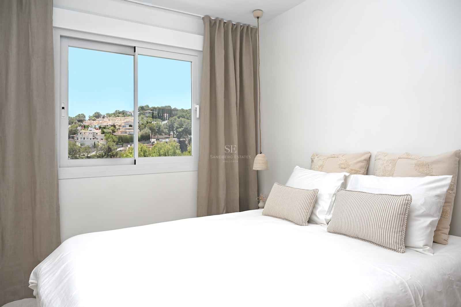 Dormitorio moderno con ropa de cama blanca, cortinas color topo y ventana con vistas a un pueblo mediterráneo.