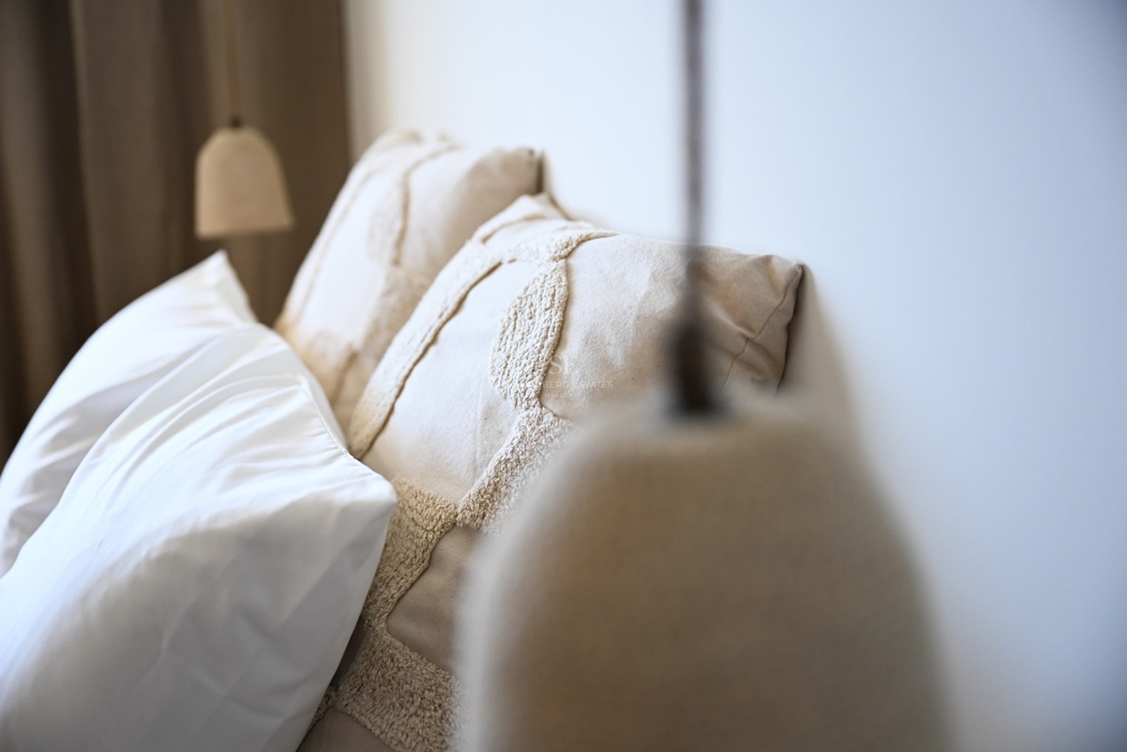 Närbild på texturerade linnekuddar och en minimalistisk taklampa i ett ljust sovrum.