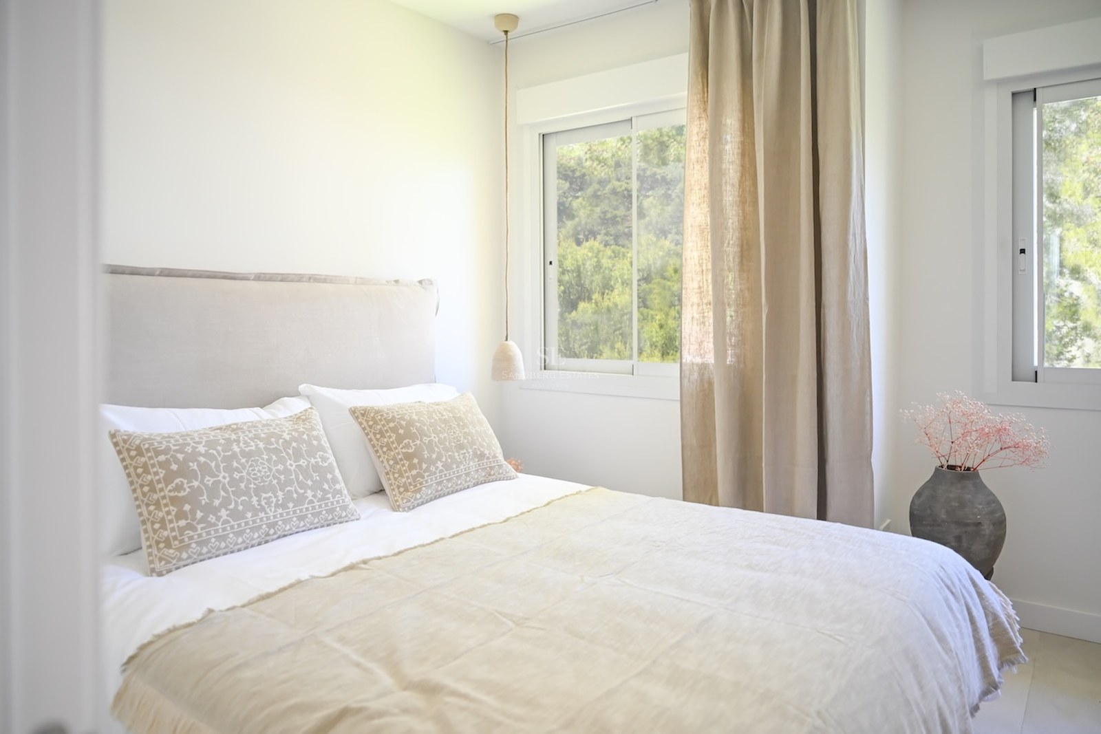 Dormitorio luminoso con cama doble, cabecero tapizado, cortinas de lino beige y vistas al jardín mediterráneo.