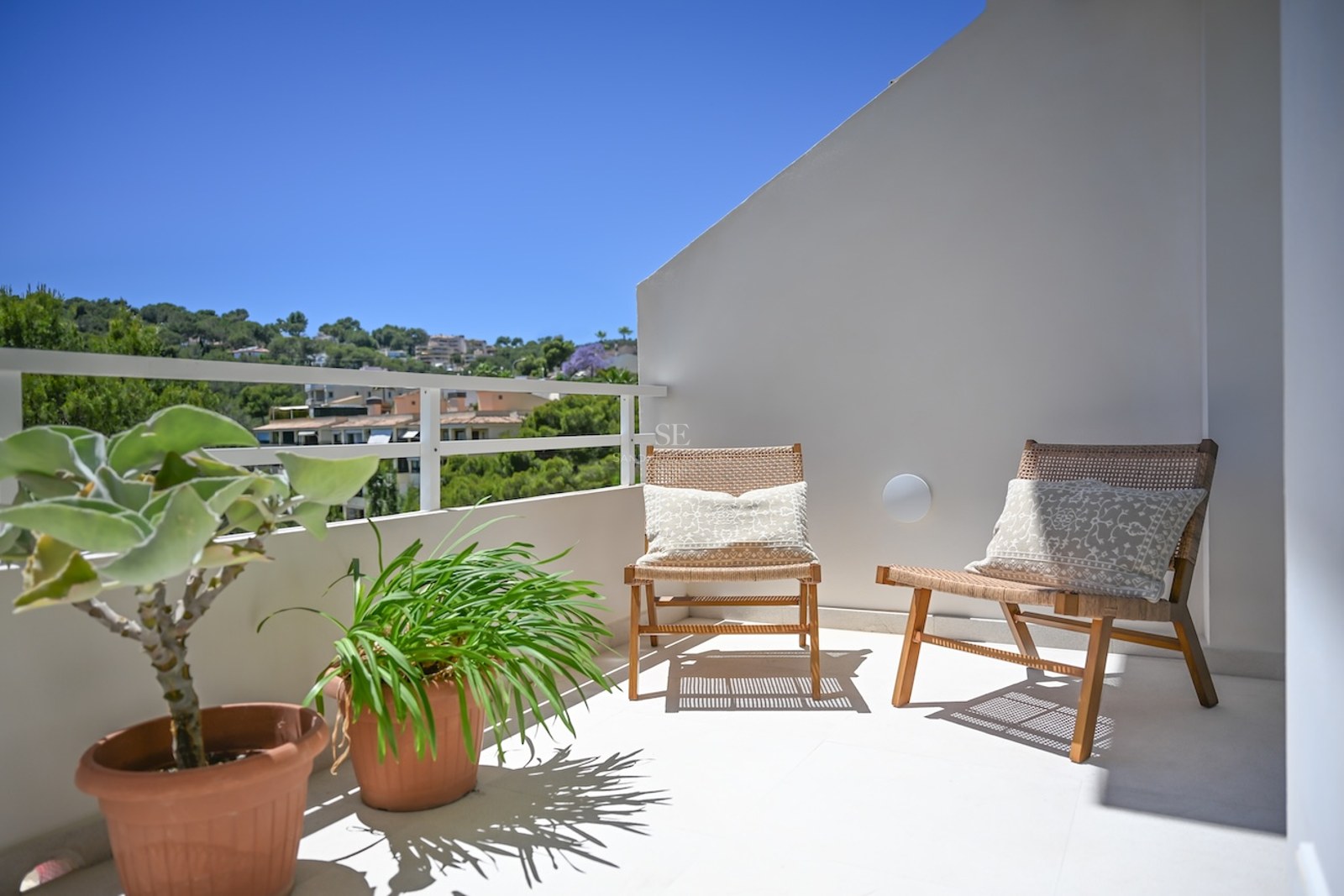 Dos sillas de mimbre en una terraza blanca y soleada con vistas a una colina mediterránea verde bajo un cielo azul.