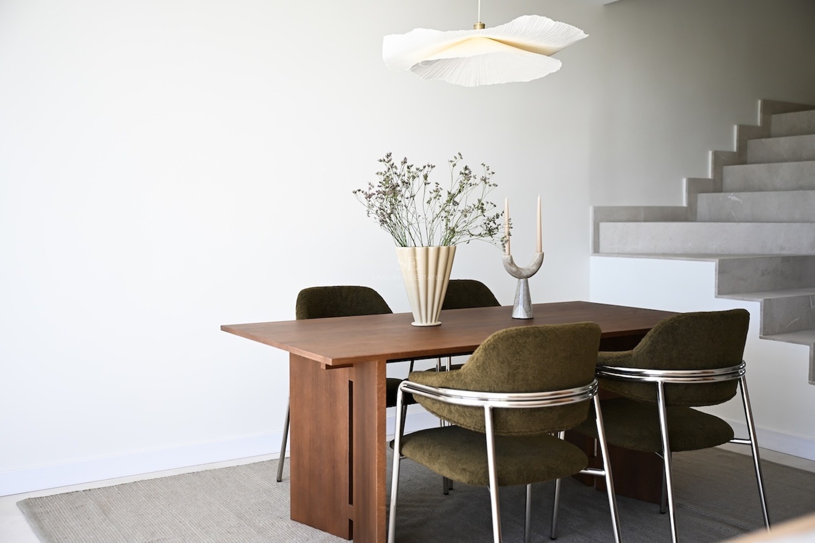 Mesa de comedor de madera con sillas verde oliva y lámpara de diseño frente a una escalera de piedra blanca.