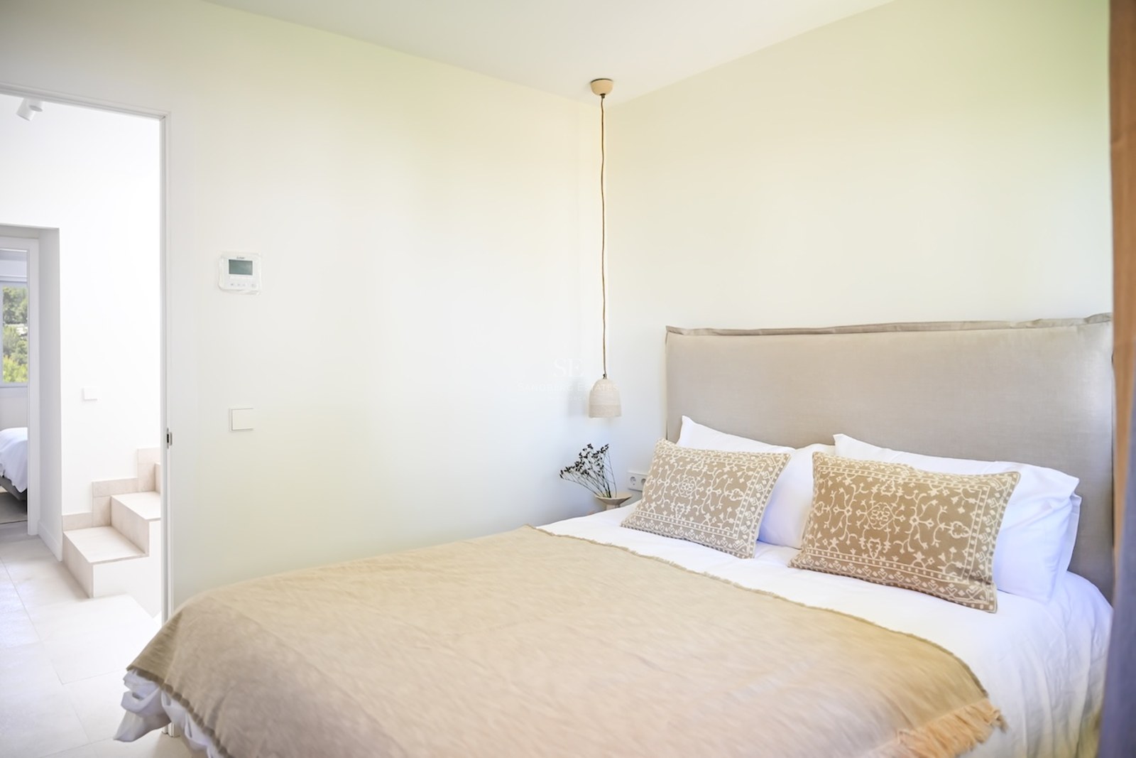Dormitorio minimalista con textiles neutros, cabecero acolchado y lámpara colgante junto a paredes blancas.