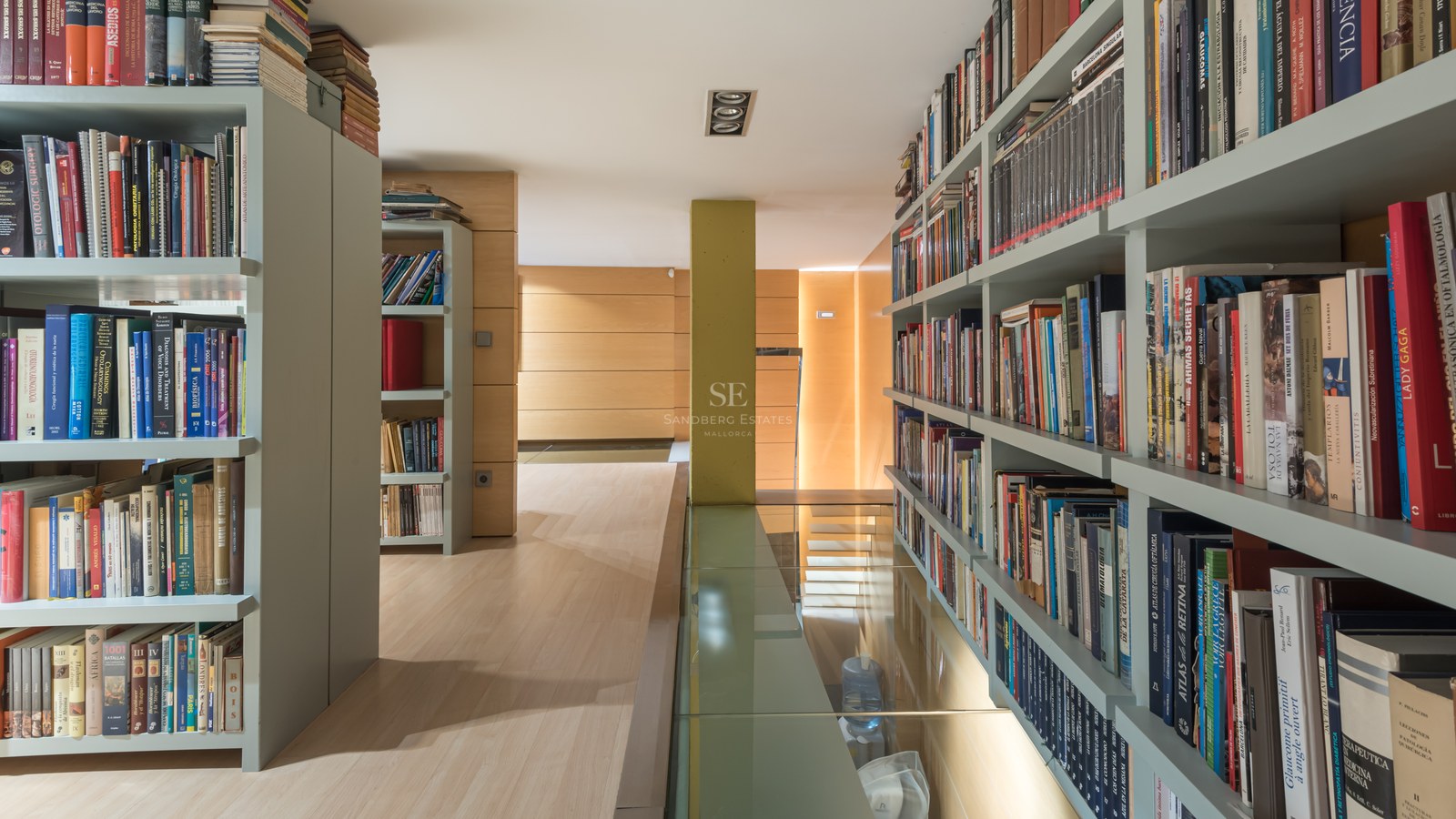 Modern home library with sage green floor-to-ceiling shelving, light wood floors, and a transparent glass floor panel.
