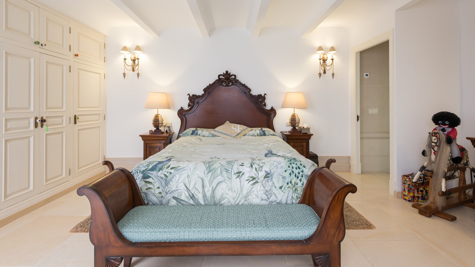 Chambre avec lit en bois sculpté, literie florale, banc au pied du lit et armoires encastrées blanches.