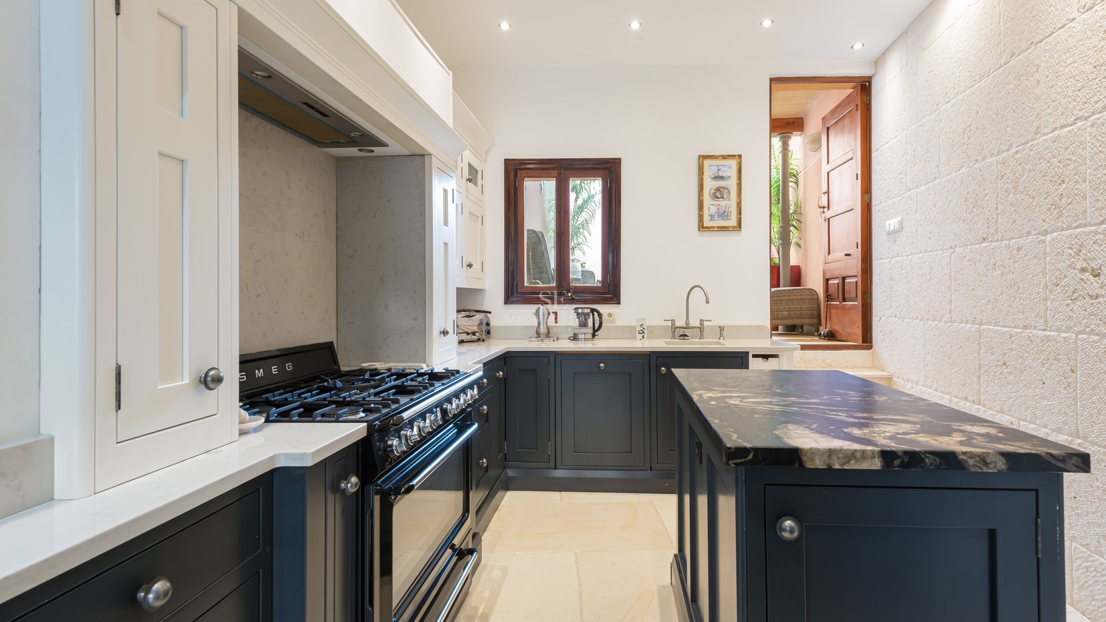 Cuisine avec des meubles bleu marine, des plans de travail en marbre blanc, une cuisinière Smeg et un mur en pierre.