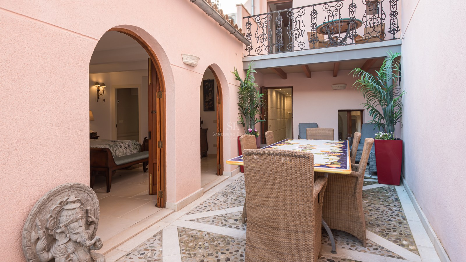 Patio privé avec sol en mosaïque de galets, mobilier en osier et murs roses aux arcades élégantes.