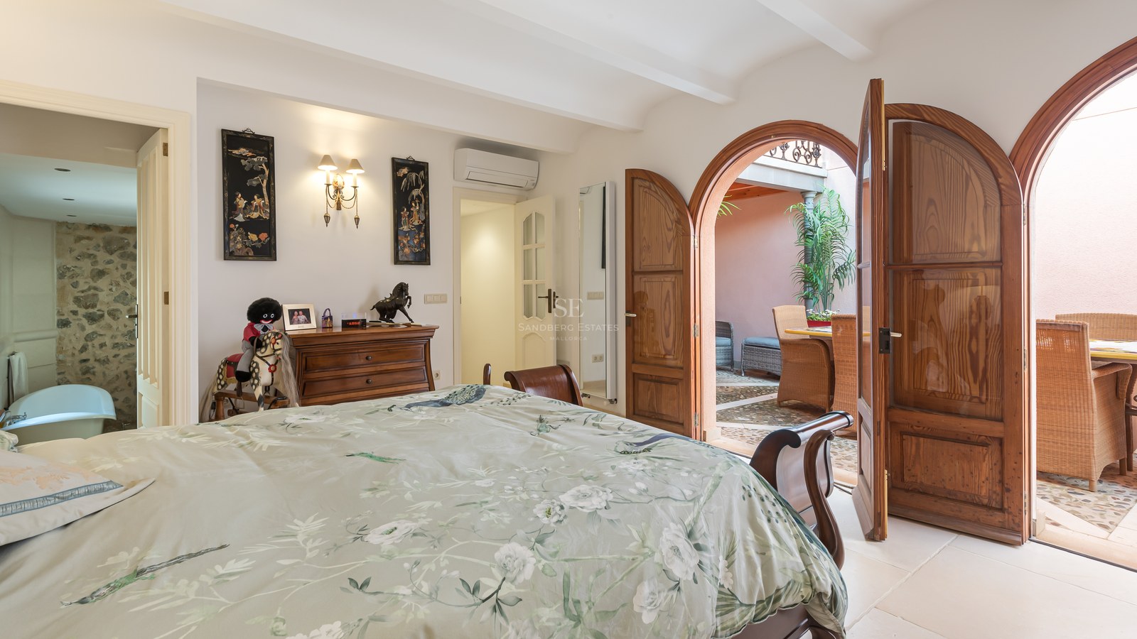 Chambre avec portes en bois voûtées ouvrant sur un patio ensoleillé, comprenant une salle de bain avec mur en pierre.