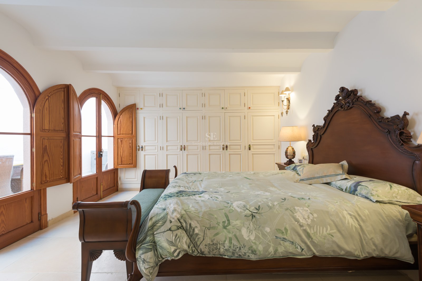 Grande chambre avec lit en bois sculpté, literie florale, placards blancs et portes cintrées en bois avec volets.
