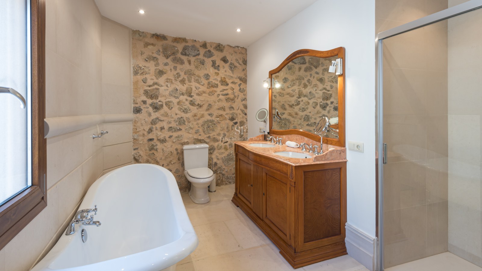 Salle de bain avec mur en pierre, double vasque en bois, baignoire îlot et douche vitrée.