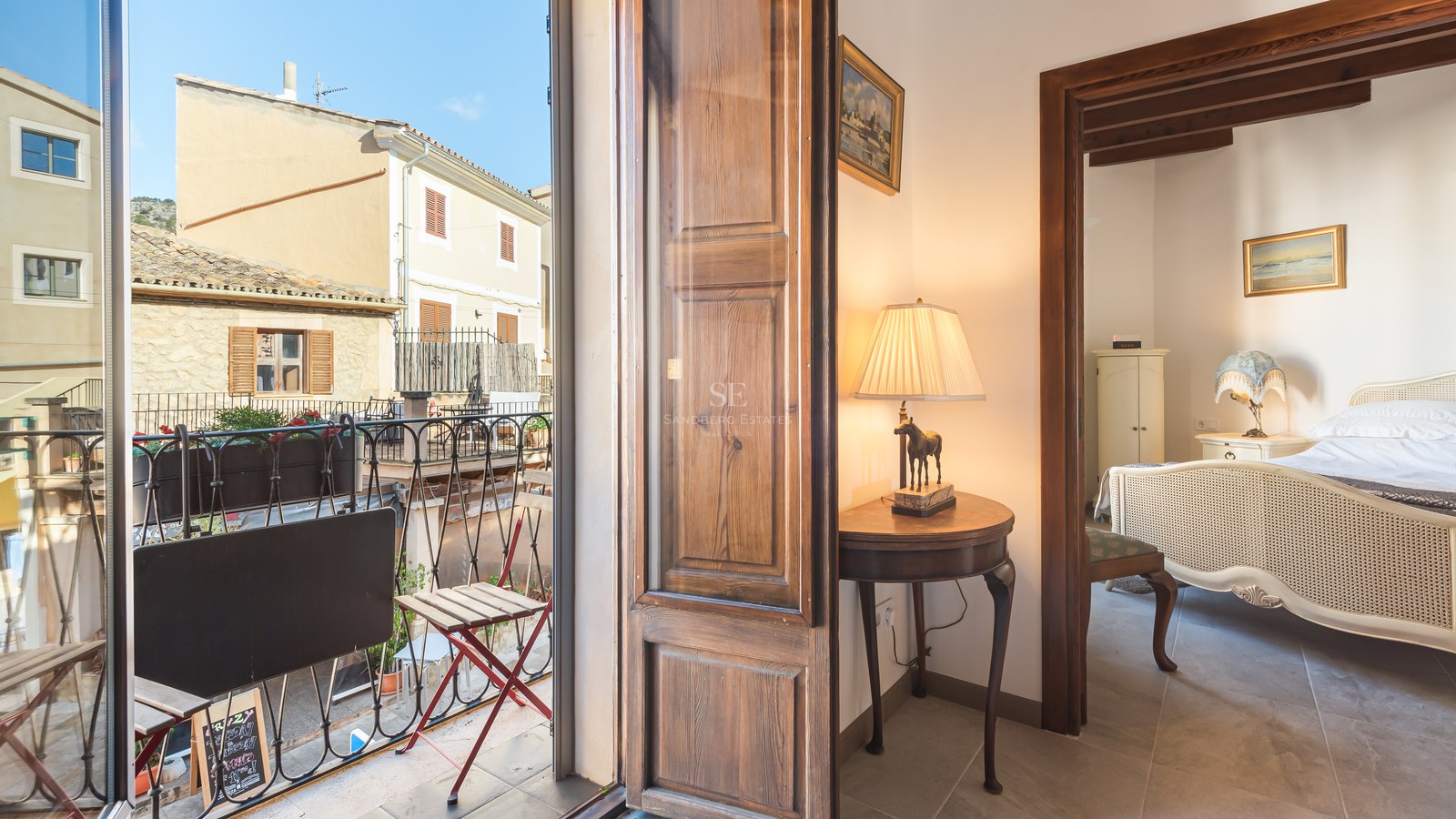 Chambre classique avec lit orné vue depuis un couloir à côté d'un balcon donnant sur une rue méditerranéenne.