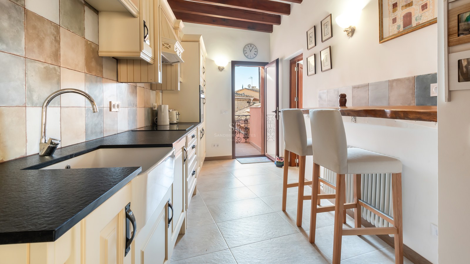 Cuisine avec plan de travail sombre, évier de ferme, poutres en bois et accès à une terrasse ensoleillée.