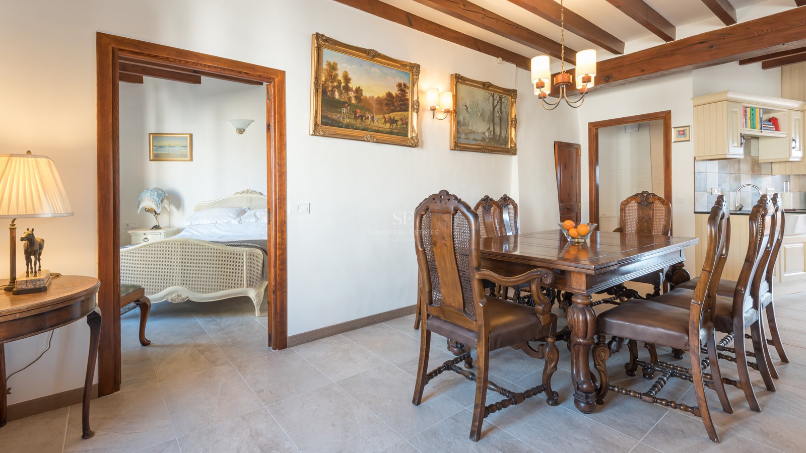 Salle à manger avec table en bois sculpté, chaises en cuir et rotin, plafond à poutres et vue sur une chambre.