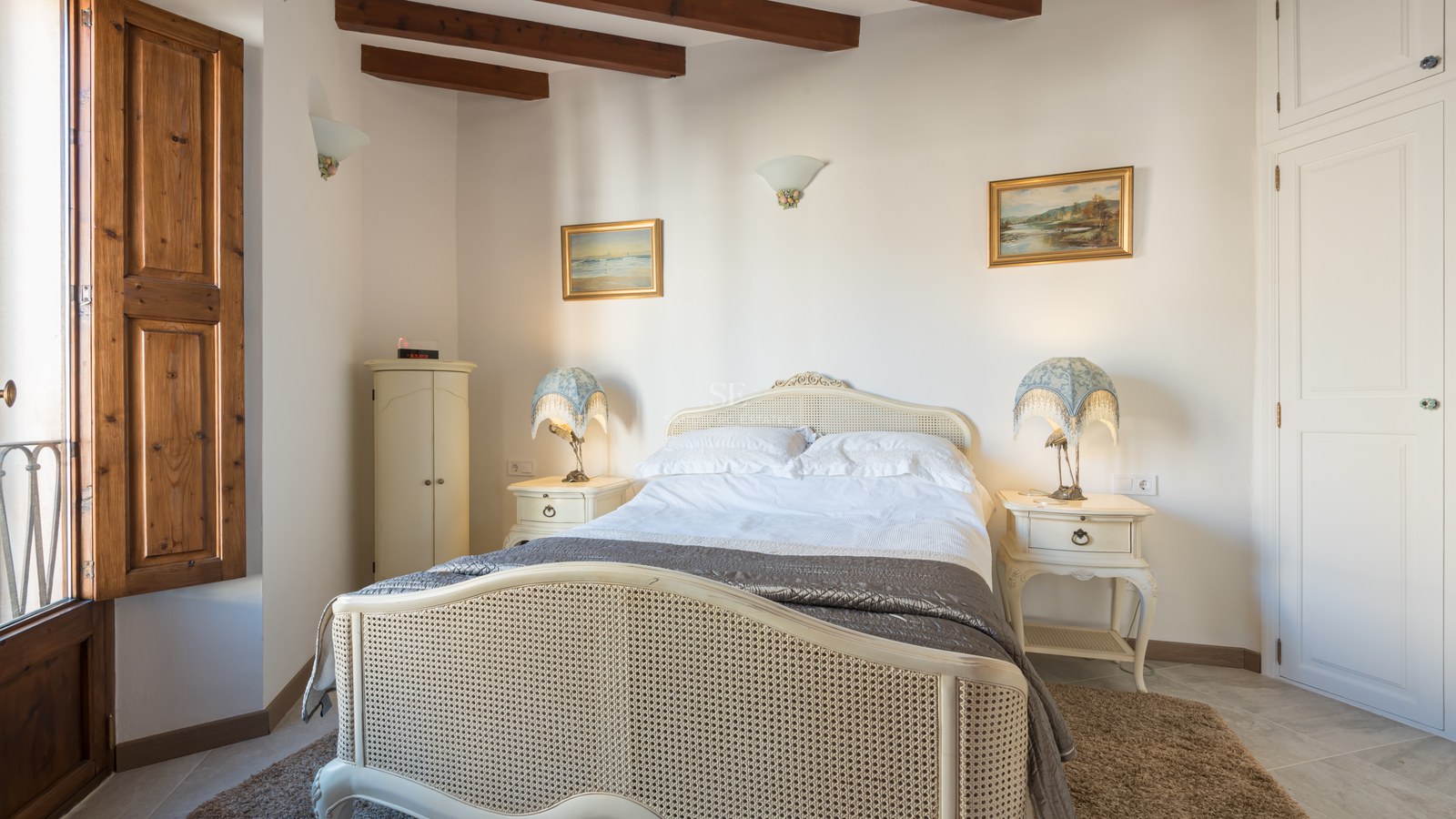Chambre chaleureuse avec lit en rotin, poutres en bois au plafond et volets intérieurs traditionnels.
