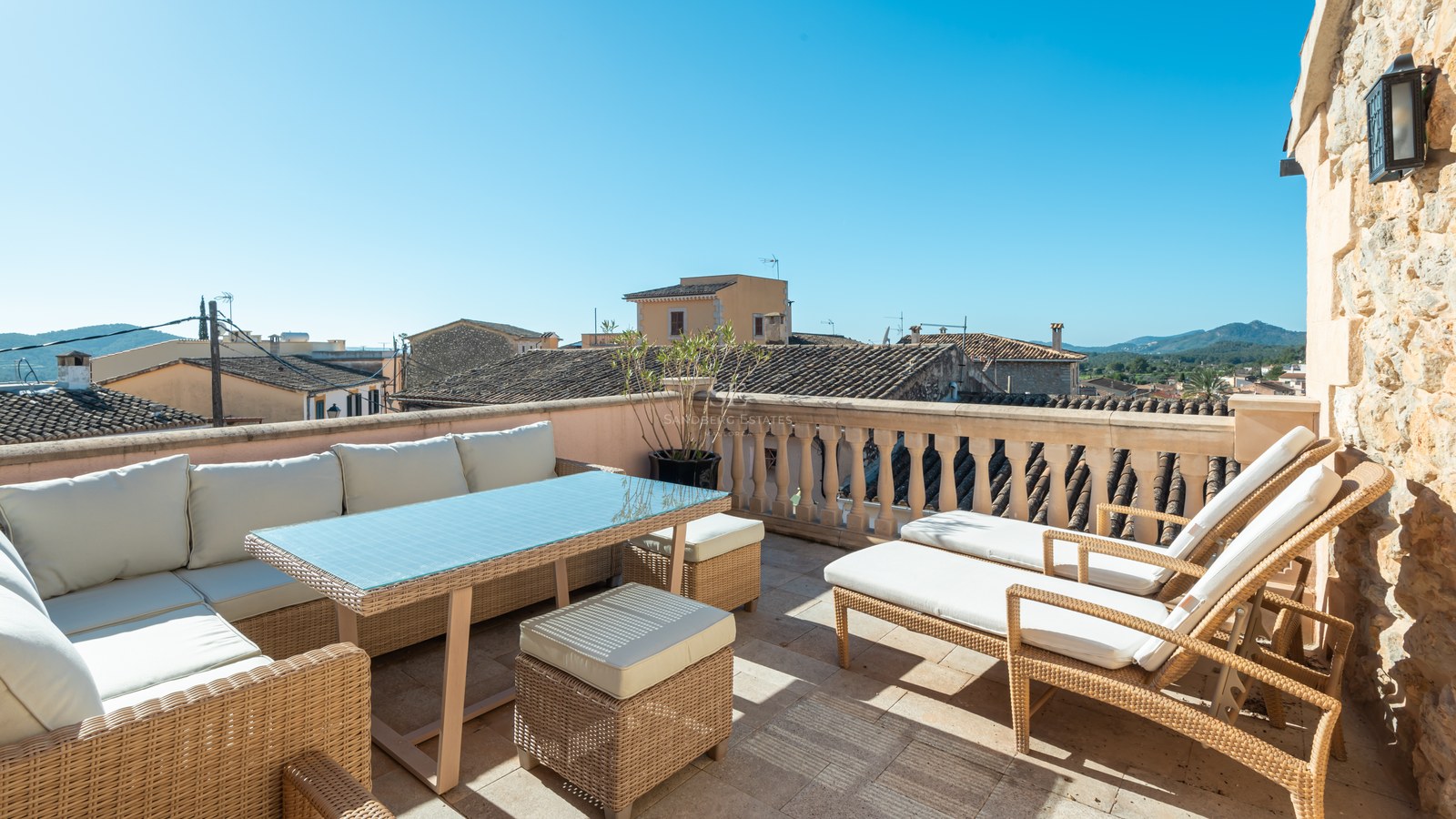 Terrasse ensoleillée avec mobilier en rotin, coussins blancs et vue sur les toits historiques et les collines.