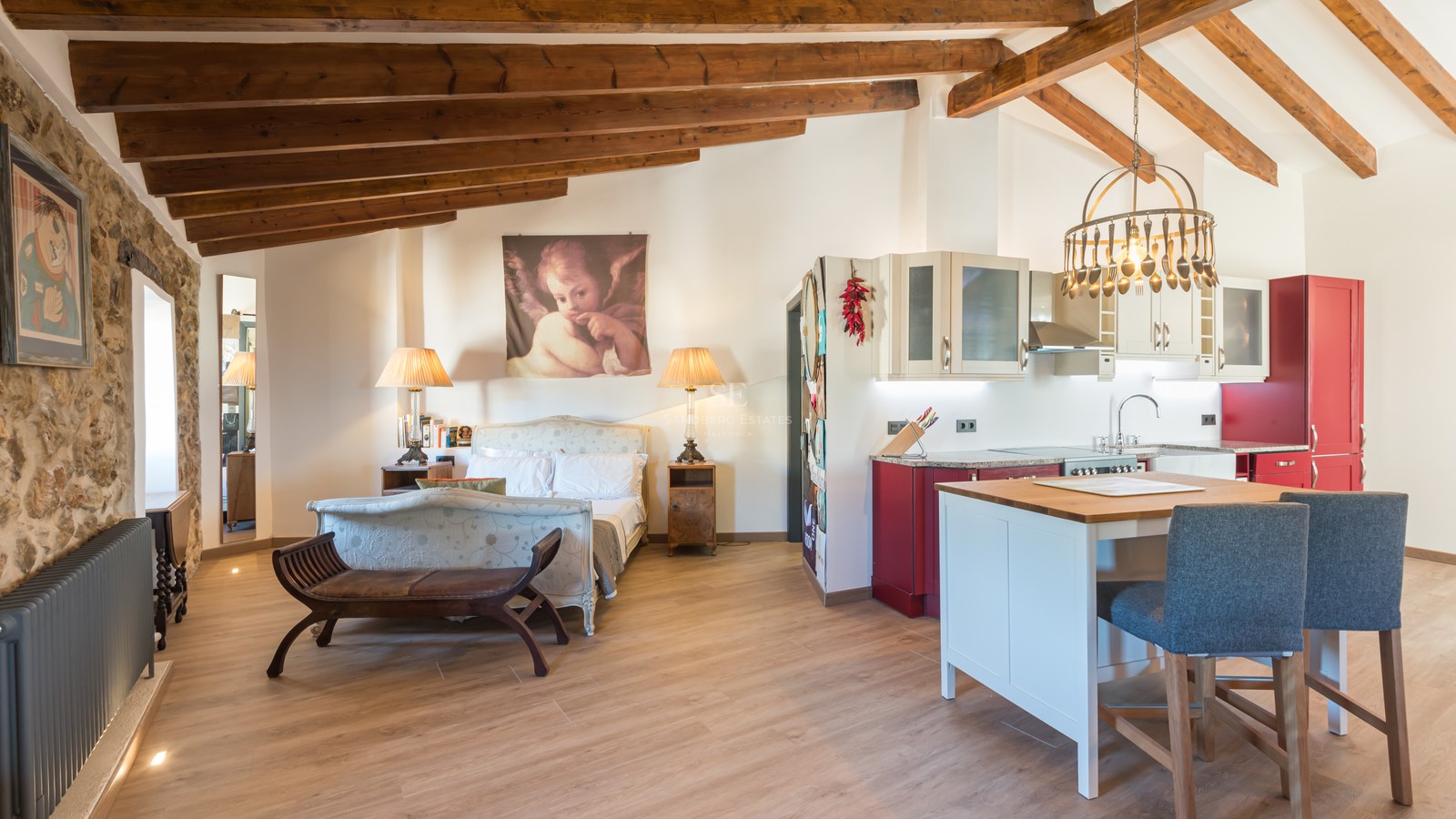 Studio avec lit classique, poutres en bois apparentes, mur en pierre et cuisine rouge et blanche avec îlot.