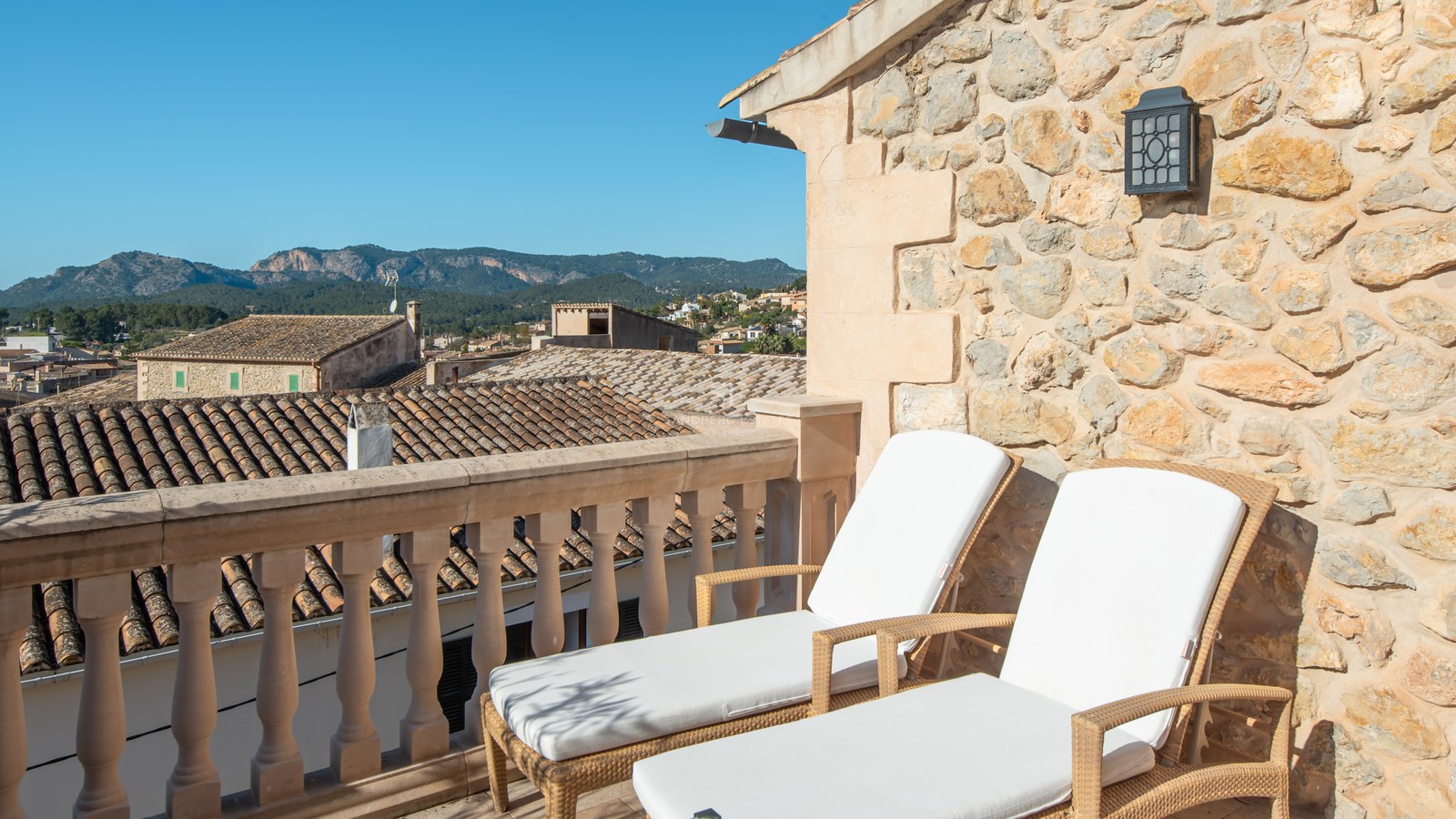 Deux chaises longues blanches sur une terrasse en pierre avec vue sur un village méditerranéen et les montagnes.