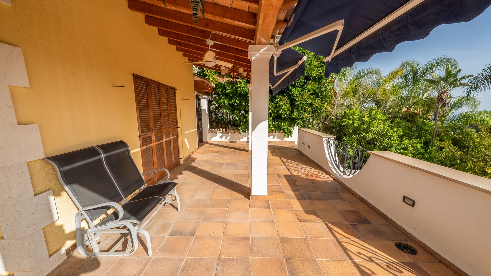 Sonnige Terrasse mit Terrakottafliesen, Holzbalken an der Decke, Deckenventilator und blauer Markise.