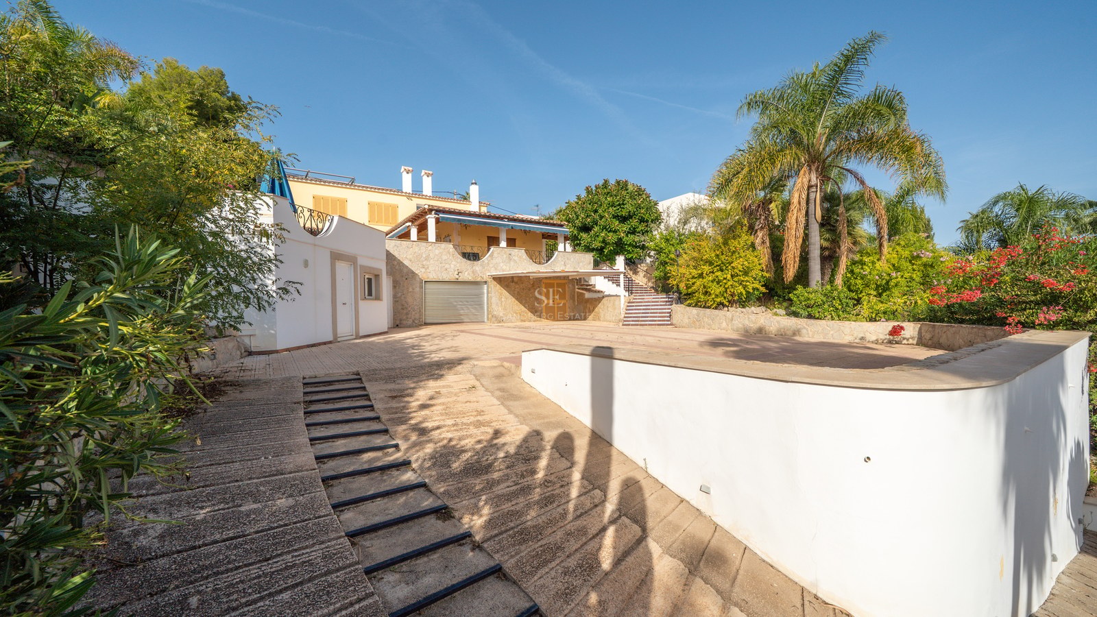 Gepflasterte Einfahrt mit Garage, umgeben von mediterraner Bepflanzung und Palmen.