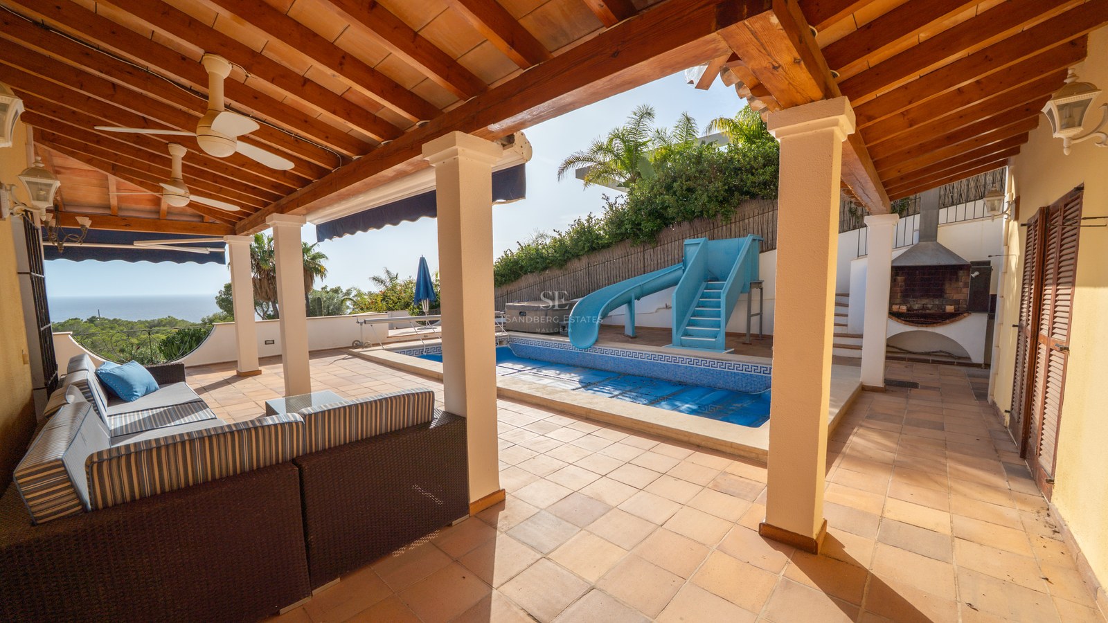 Überdachte Terrasse mit Holzbalken und Sitzgelegenheiten mit Blick auf einen blauen Pool mit Rutsche und Palmen.