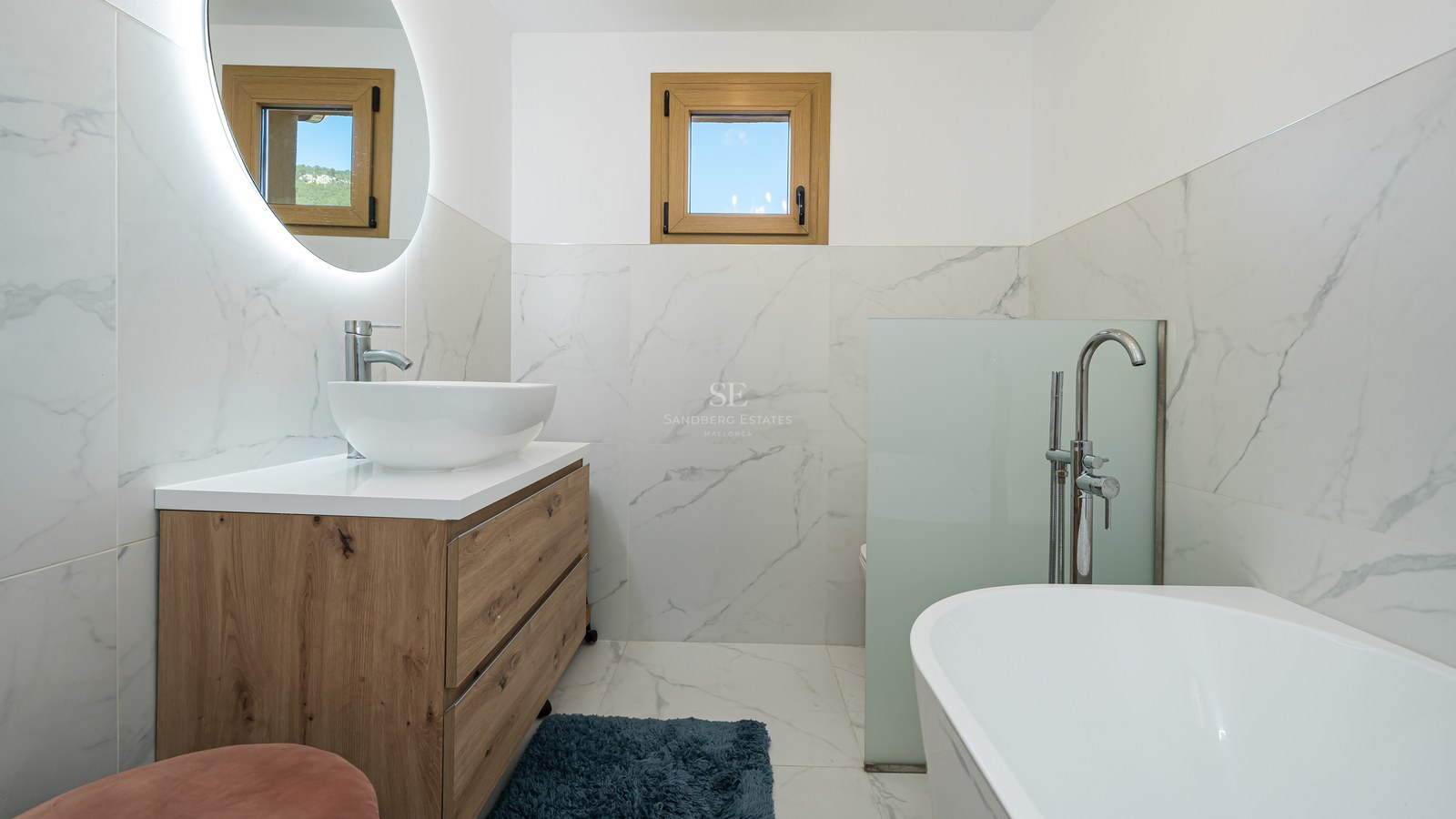 Baño moderno con paredes de mármol, mueble de madera, lavabo de sobremesa, espejo retroiluminado y bañera blanca.