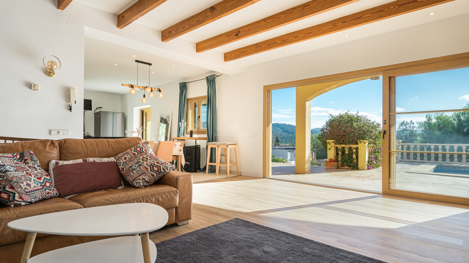 Salón con sofá de cuero marrón, vigas de madera y puertas correderas que dan a una terraza con vistas a la montaña.