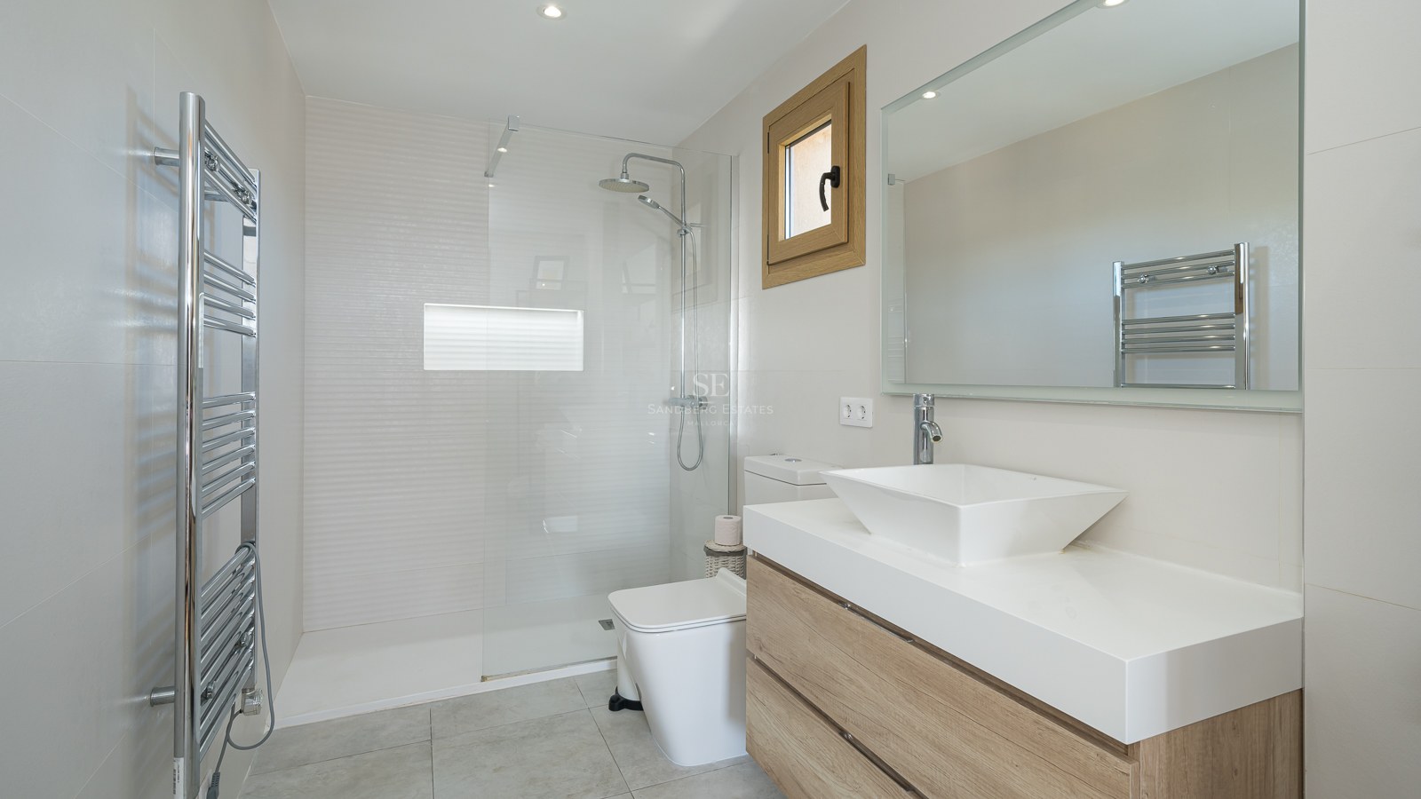 Baño moderno con ducha de cristal, mueble de madera con lavabo cuadrado y toallero radiador cromado.