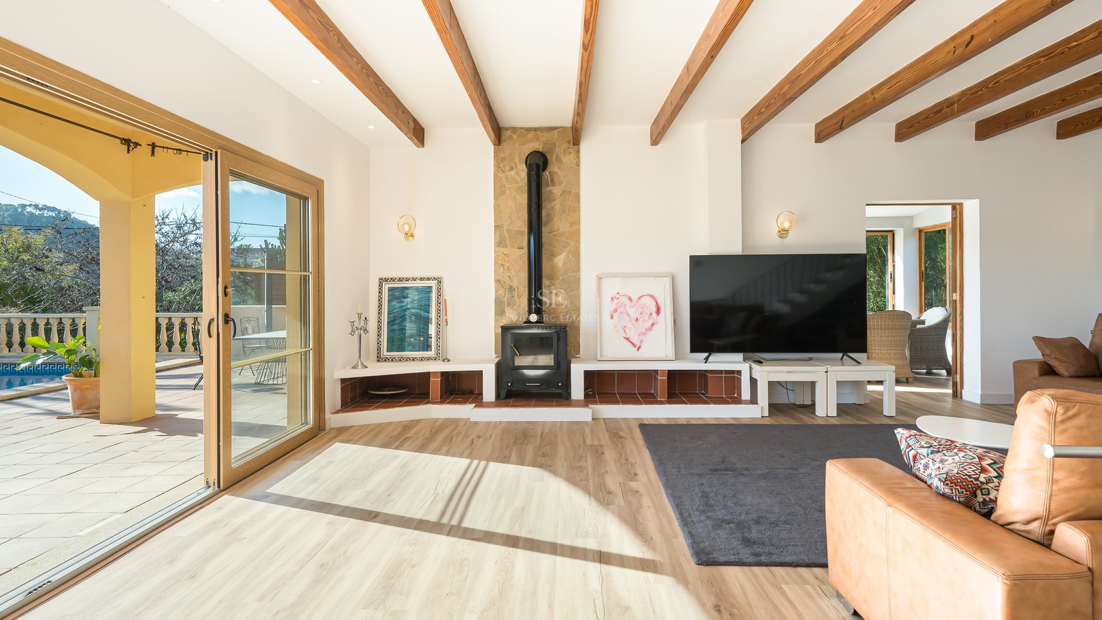 Salón moderno con vigas de madera, chimenea de leña y puertas correderas que dan a una terraza soleada.