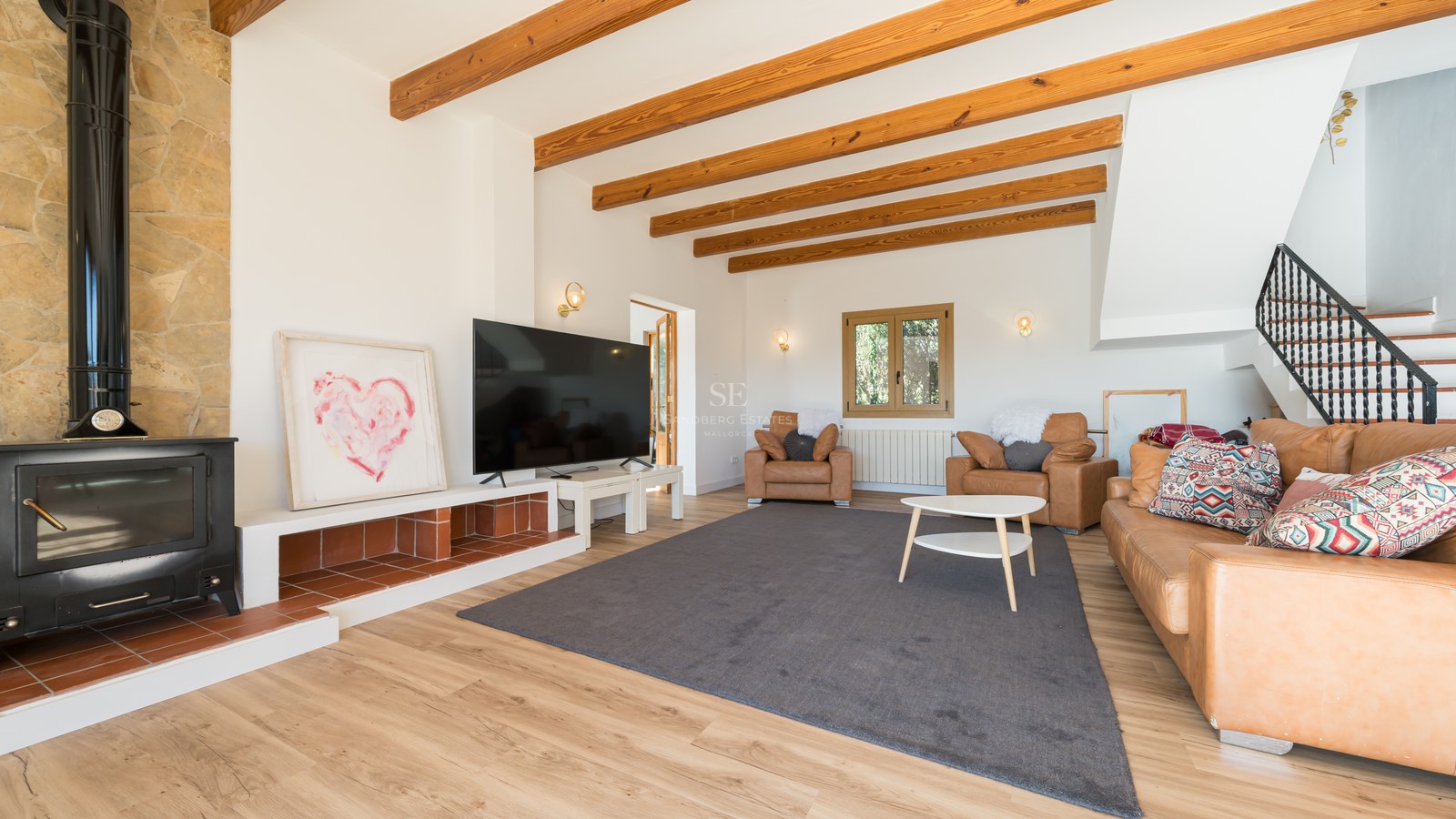 Acogedor salón con vigas de madera a la vista, estufa de leña negra, sofás de cuero y una televisión grande.