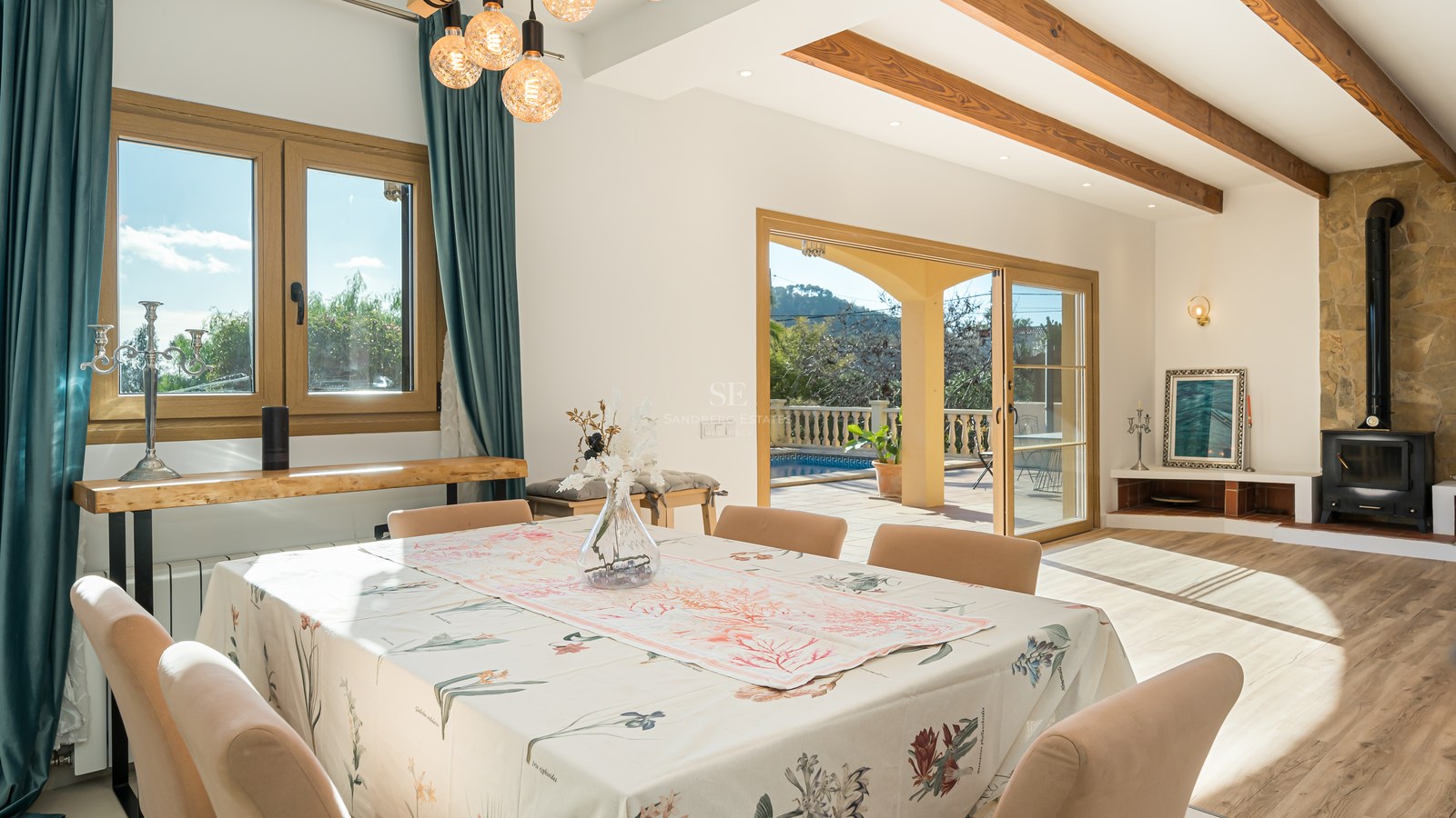 Comedor luminoso con vigas de madera, chimenea de piedra y puertas de cristal hacia una terraza con piscina.