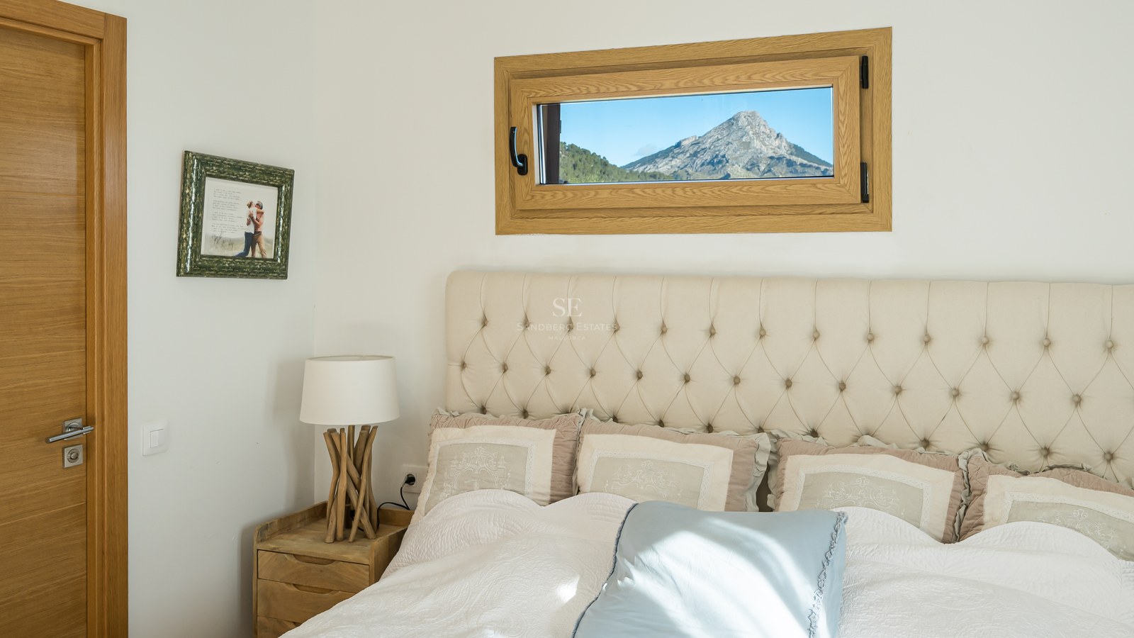 Cama con cabecero capitoné beige junto a una ventana pequeña que enmarca una montaña escarpada.