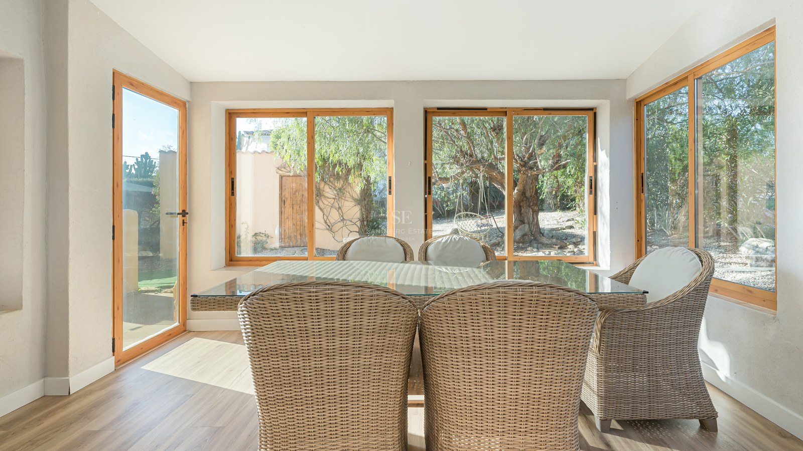 Comedor interior soleado con sillas de mimbre y mesa de cristal con vistas a un jardín con árboles mediterráneos.