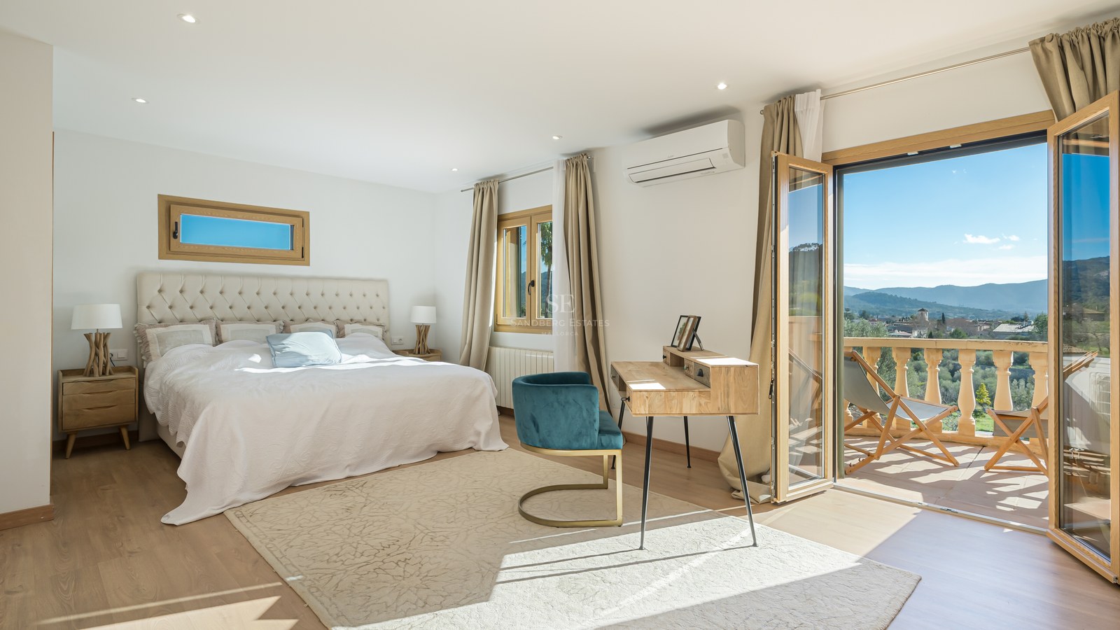 Dormitorio principal luminoso con cama grande, escritorio de madera y balcón con vistas a las montañas.