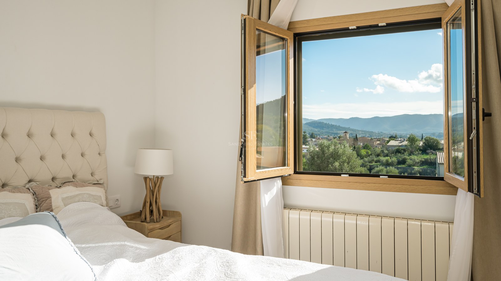 Dormitorio acogedor con cama de cabecero acolchado y ventana abierta con vistas panorámicas a un pueblo y montañas.