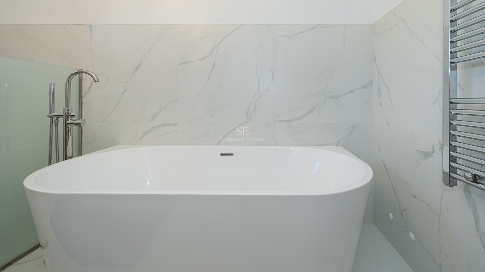 Bañera exenta blanca con grifería cromada frente a paredes de mármol con veteado gris y un radiador toallero.