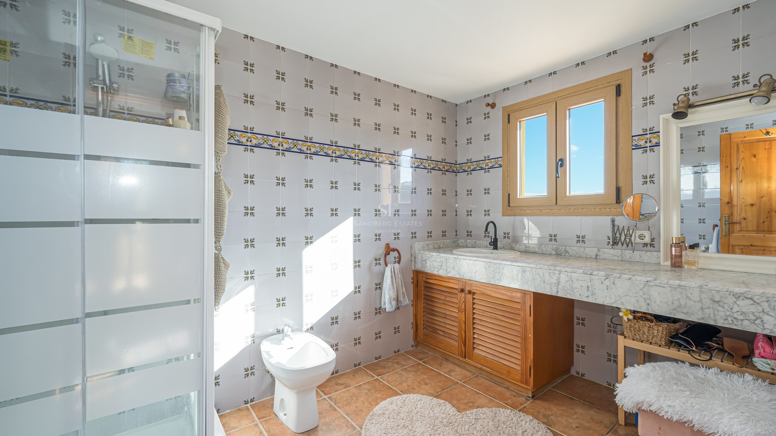 Baño con azulejos decorados, encimera de mármol blanco, mueble de madera, bidé y suelo de terracota bajo luz natural.