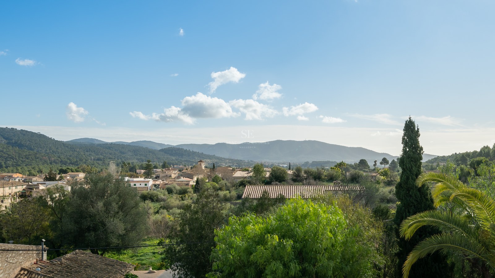 Vista elevada de un pueblo mediterráneo con tejados de terracota rodeado de vegetación y montañas verdes bajo cielo azul.