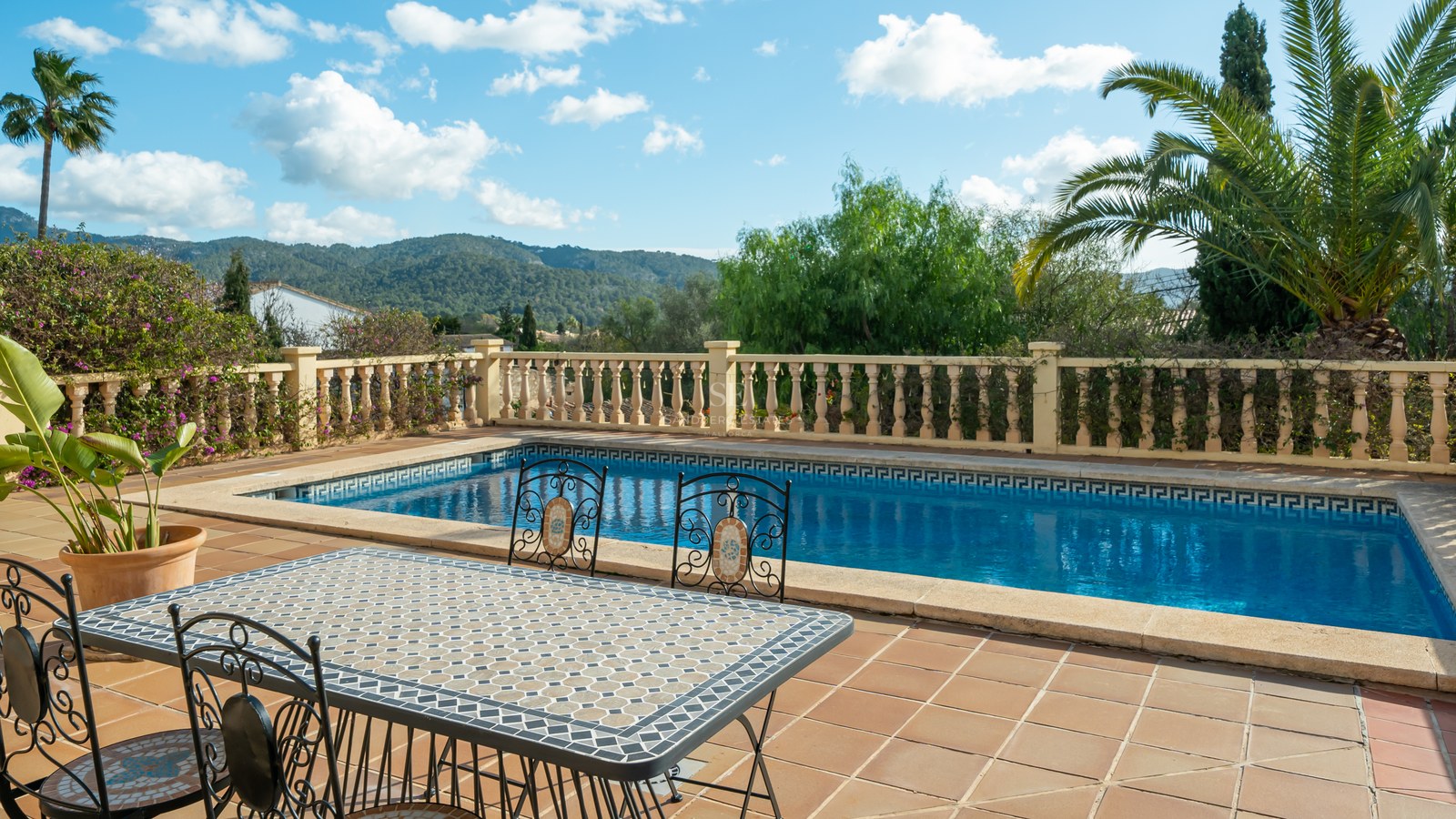 Piscina rectangular y mesa de mosaico en una terraza de terracota con vistas a la montaña y palmeras.