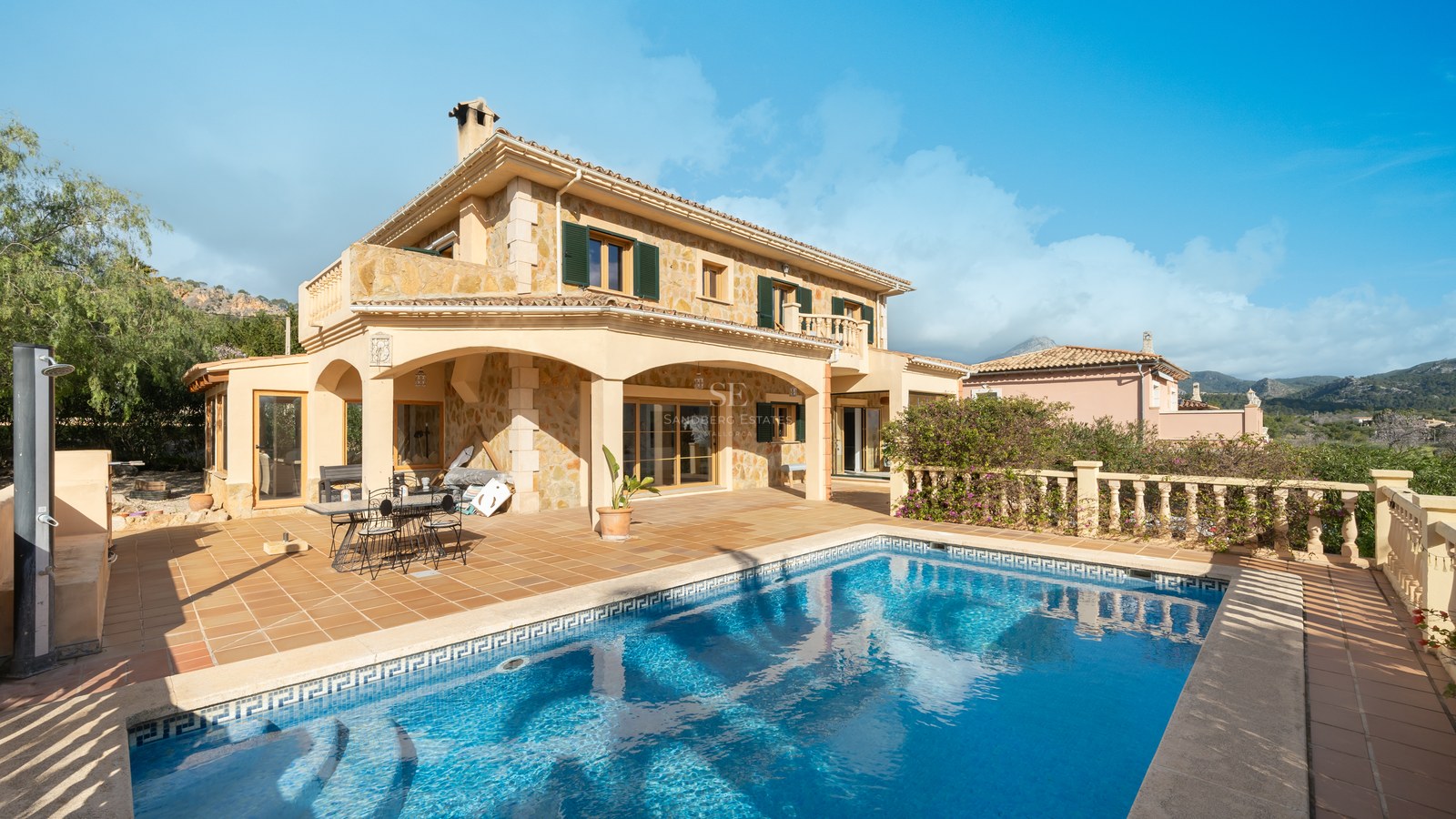 Vista de una villa de piedra de dos plantas con piscina color turquesa y terraza de terracota bajo un cielo azul.