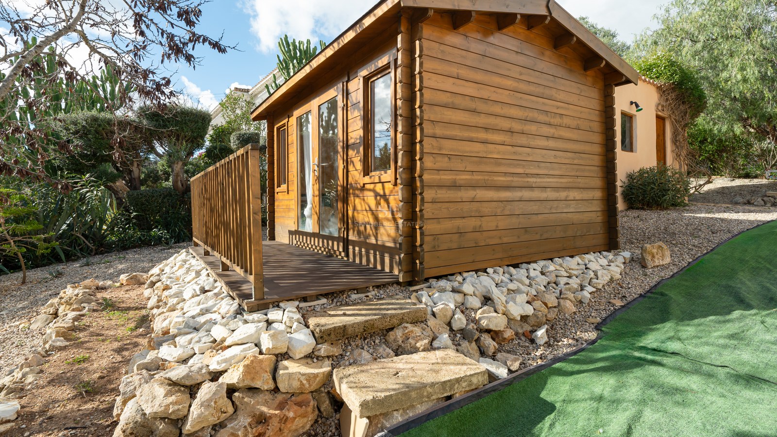 Cabaña de troncos de madera con terraza pequeña en un jardín mediterráneo con muros de piedra natural.