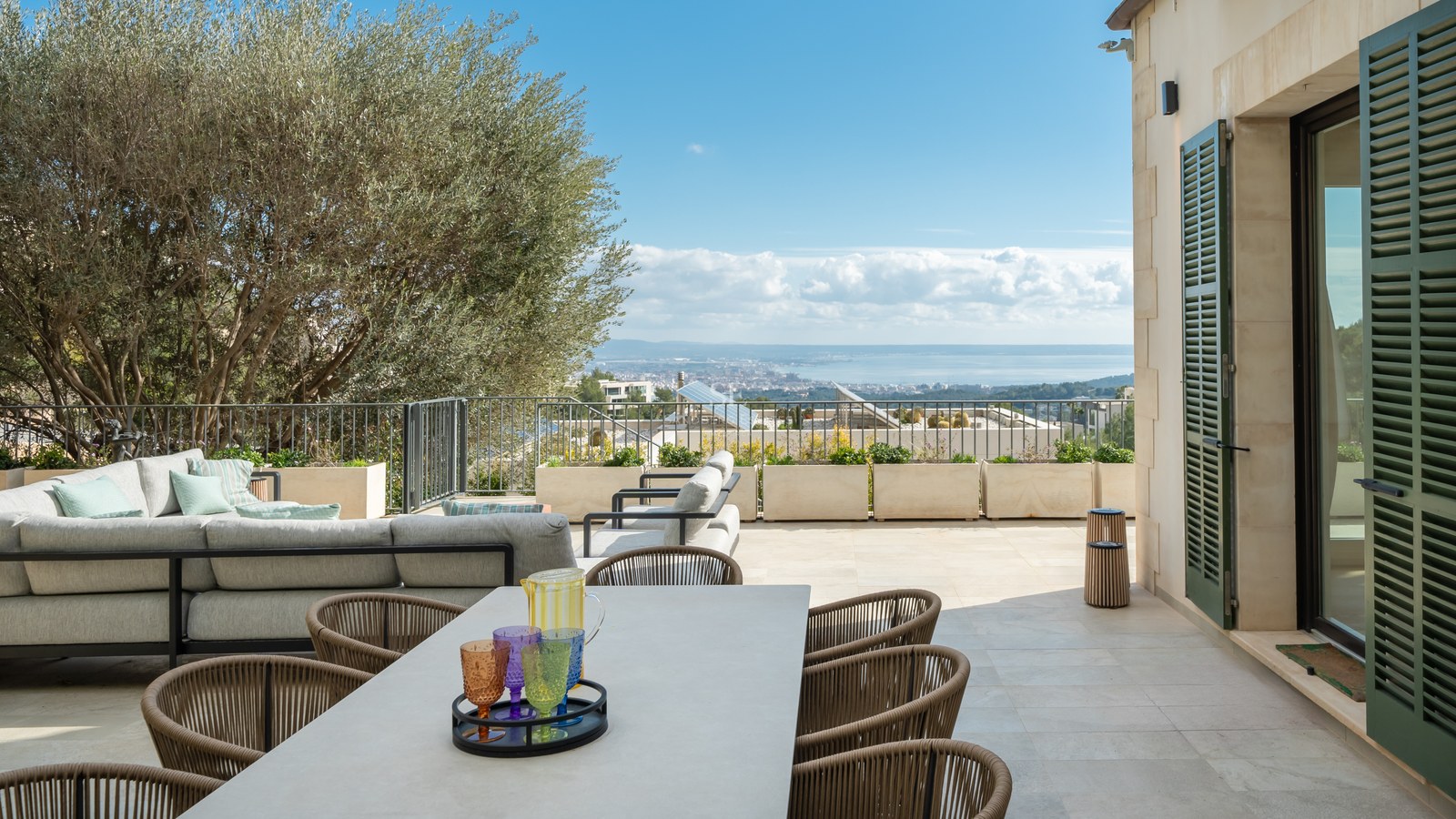 Terraza de piedra con comedor, sofás y olivo con vistas al mar y a la ciudad bajo un cielo despejado.