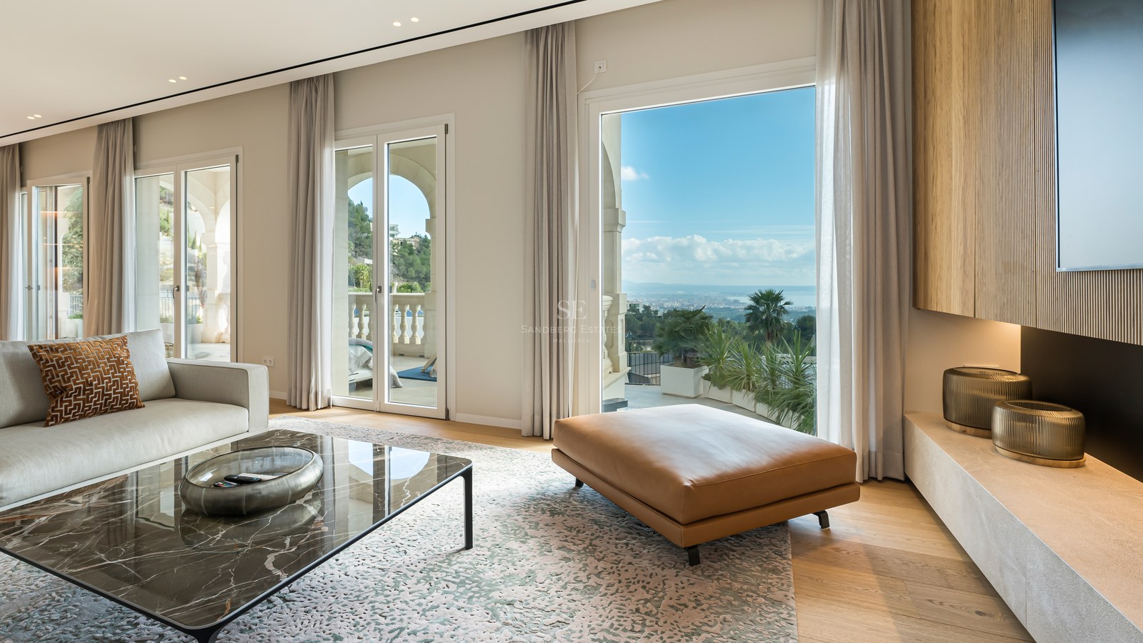 Salón moderno con mesa de mármol, otomana de cuero y ventanales con vistas al mar Mediterráneo.