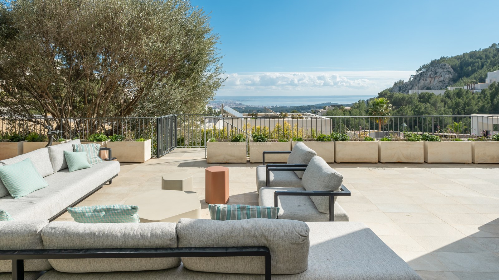Terraza moderna con sofás grises, suelo de piedra natural, un olivo y vistas despejadas al mar y la costa.