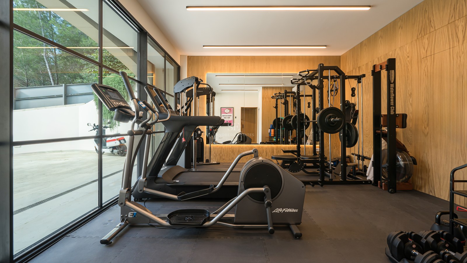 Gimnasio moderno con equipamiento de alta gama, paredes paneladas en roble y grandes ventanales.