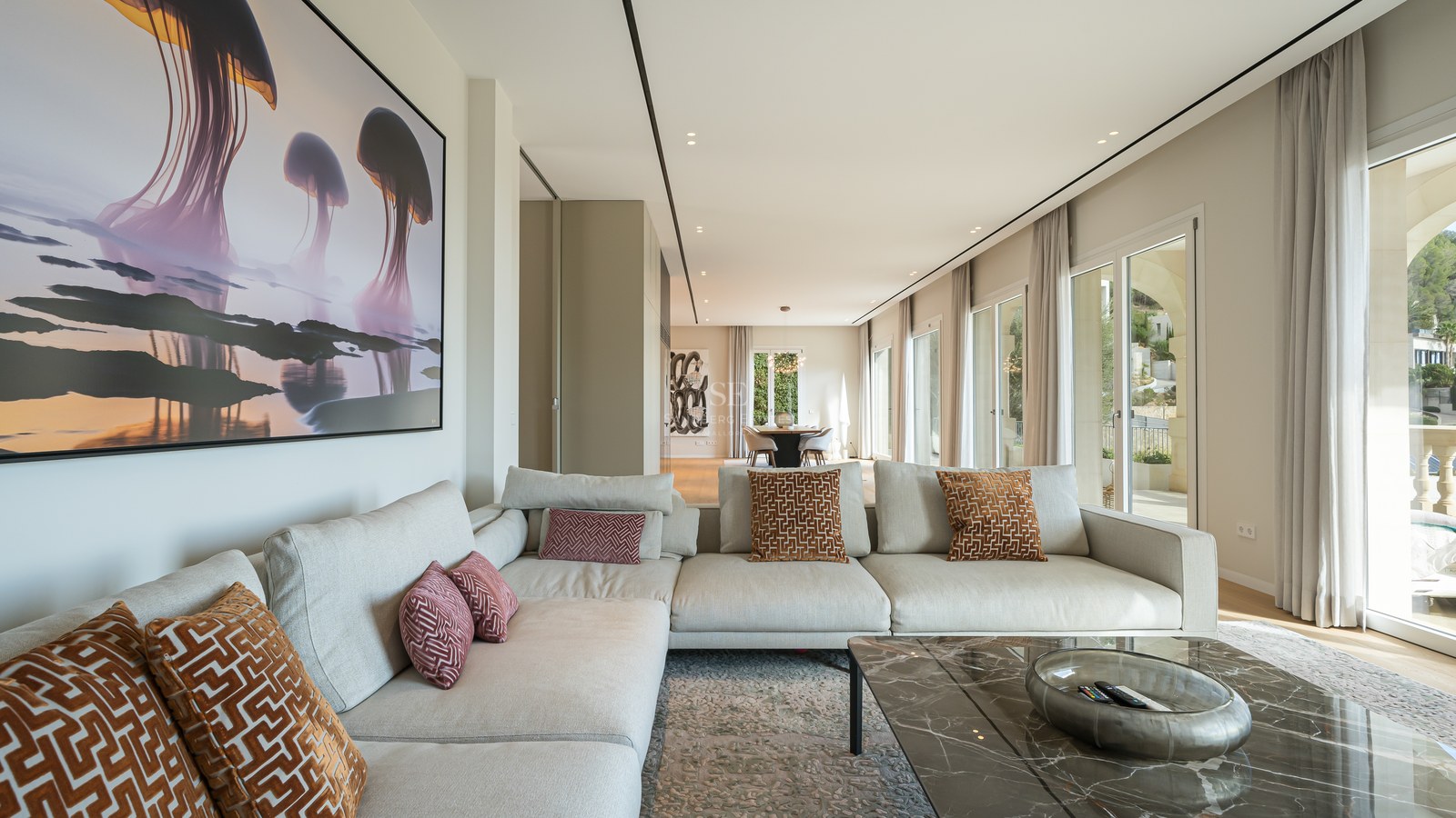 Salón moderno con sofá en L color crema, mesa de mármol y grandes ventanales con vistas al exterior.