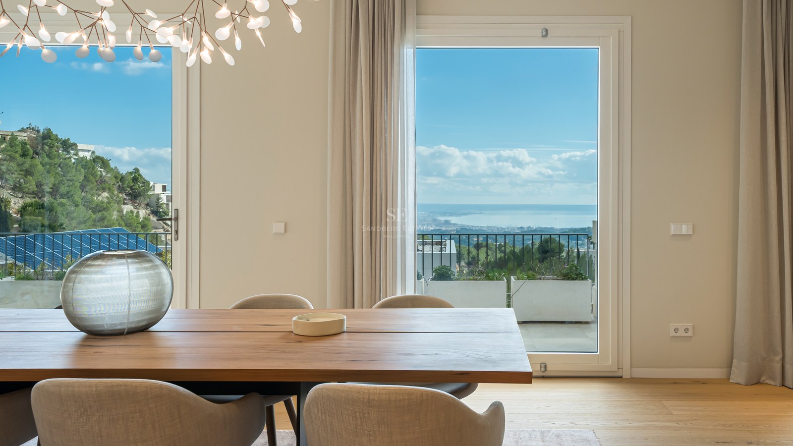 Comedor moderno con mesa de madera maciza y grandes ventanales con vistas a la costa mediterránea.