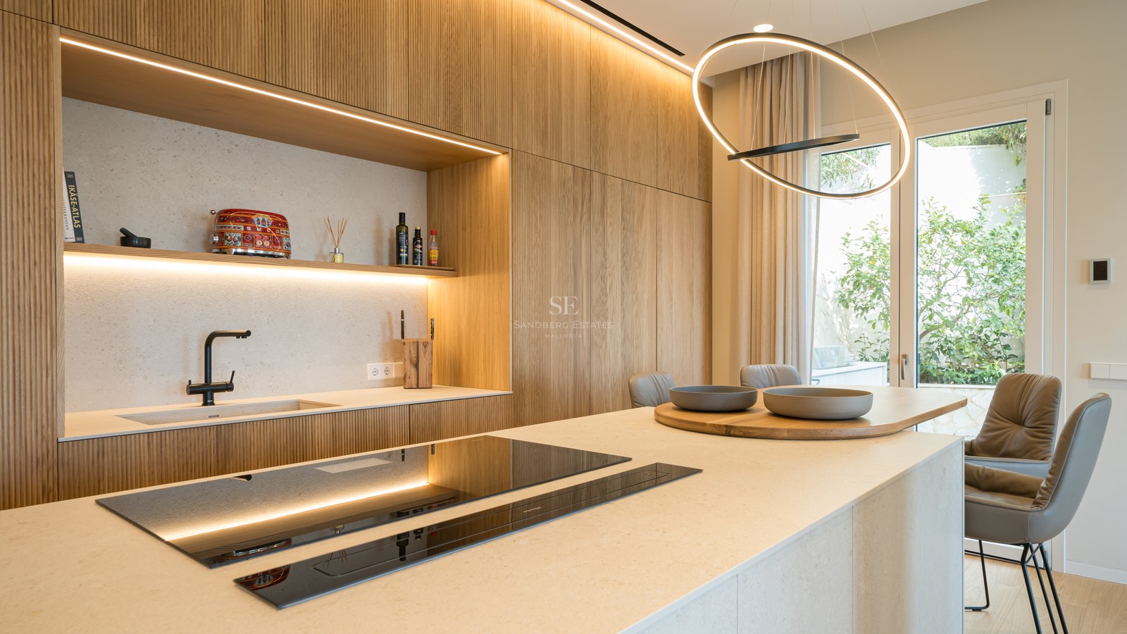 Cocina moderna con armarios de roble fresado, isla de piedra, placa de inducción y puerta de cristal al jardín.