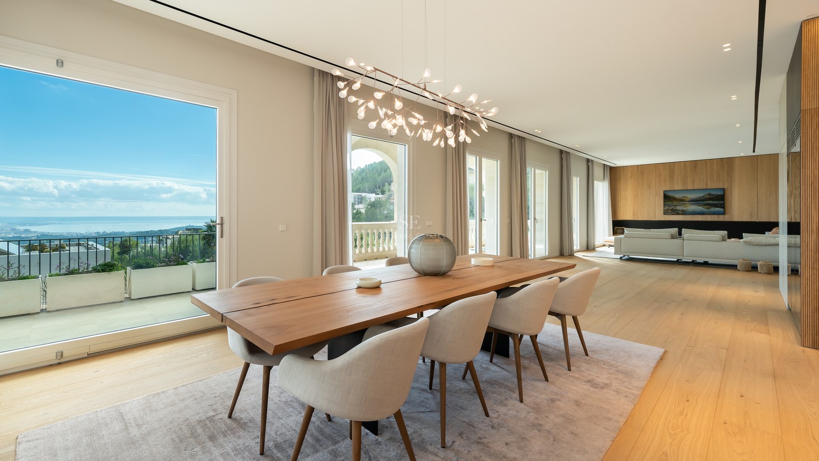 Elegante comedor con mesa de madera maciza, lámpara escultórica y grandes ventanales con vistas al mar y la costa.