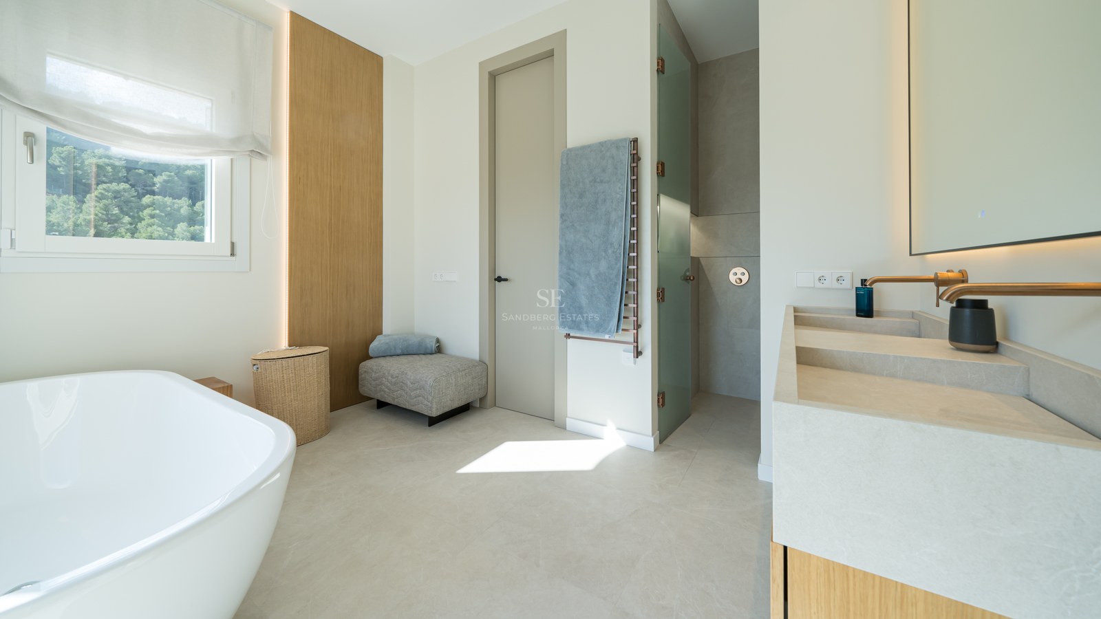 Baño moderno con bañera exenta, lavabo doble de piedra natural, grifería de bronce y vistas al bosque.